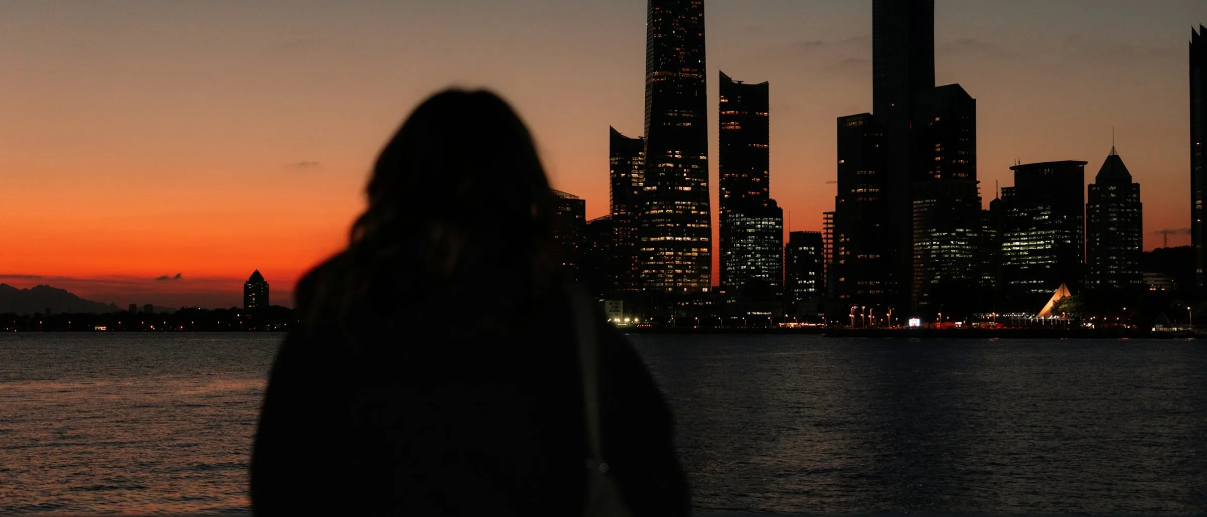Silueta de una persona de espaldas mirando el perfil de una ciudad al atardecer, con las luces encendidas a la derecha
